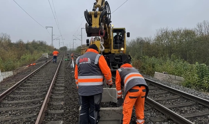 Zwei Bauarbeiter in orangefarbenen Warnkleidungen arbeiten an Gleisen vor einem gelben Gleisbagger unter grauem Himmel