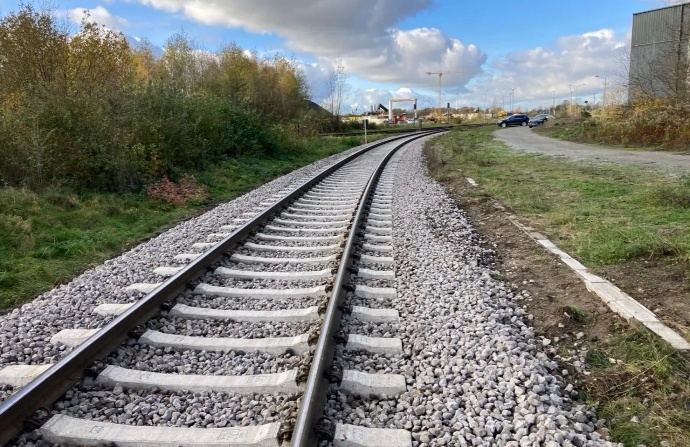 Gleise einer Eisenbahnstrecke, die sich nach rechts krümmen, mit Schotterbett und links Büschen sowie rechts einem Parkplatz mit einem blauen Auto